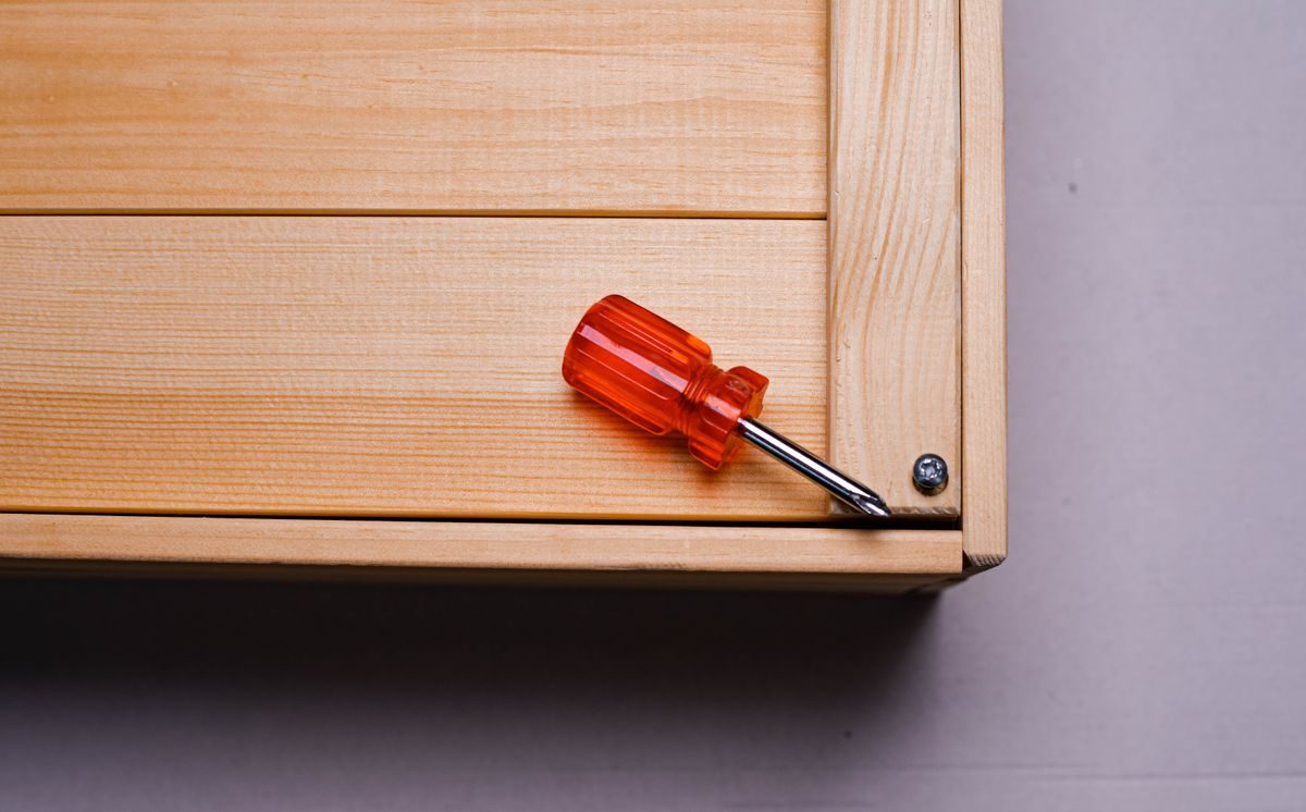 A screwdriver resting on the corner of a wooden cabinet beside a single screw